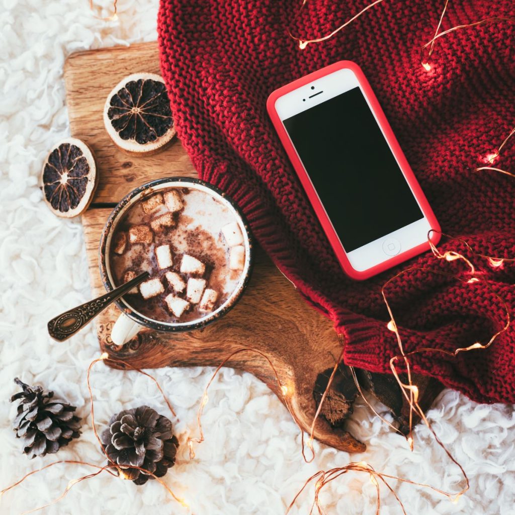 a mug of hot cocoa on a wooden platter next to dried oranges and an iphone sitting on a red knit blanket