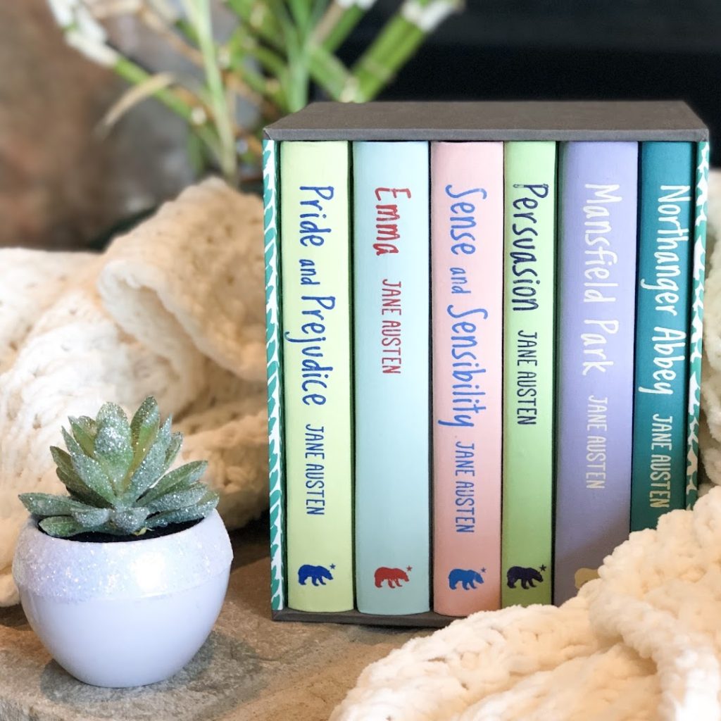 Pastel colored books next to a small houseplant and white knit blanket sitting on a stone fireplace